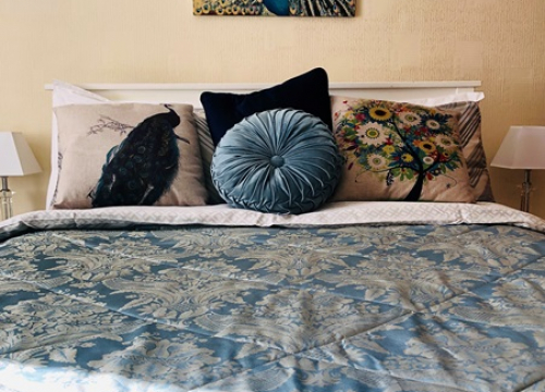 A Pendrin bed dressed in blue damask with peacock cushions beside a bright window